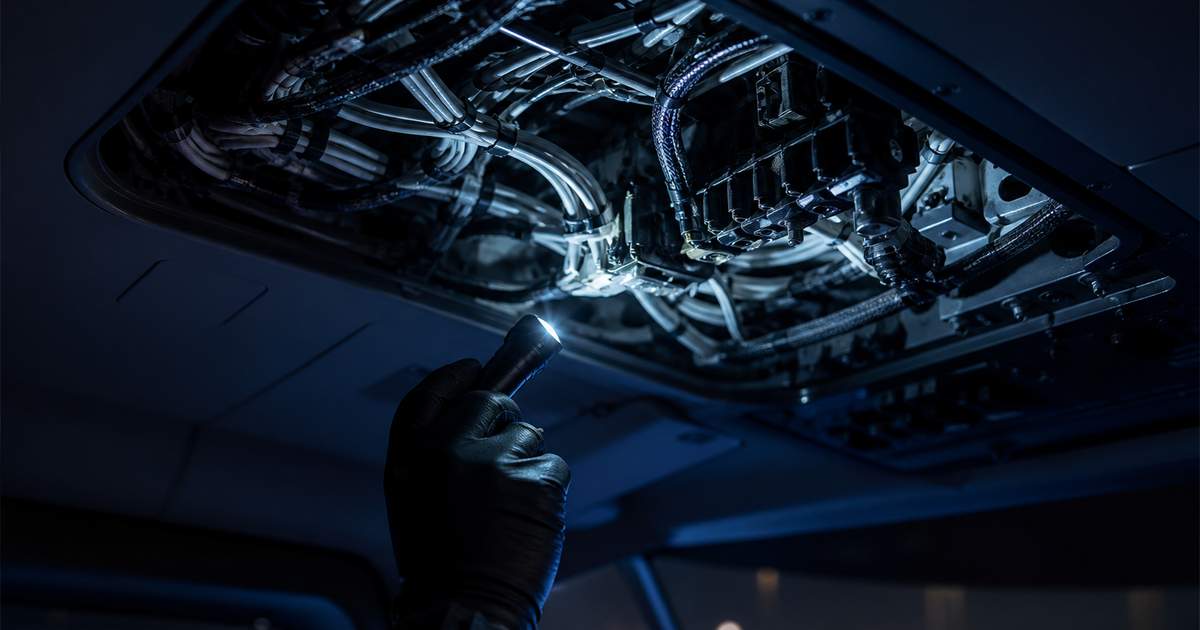 Geöffnetes Overhead-Panel eines Hubschraubercockpits mit freiliegendem Kabelbaum, Inspektionsleuchte in behandschuhter Hand